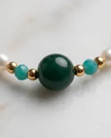Collar de acero con baño de oro, compuesto por una combinación de perlas naturales blancas, piedras semipreciosas en tonos verdes y turquesas, y pequeñas cuentas doradas. Presenta un dije central de piedra verde oscura y un cierre con cadena de alargue que incluye una medalla grabada con las iniciales MA.