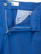 Pantalón de vestir color crema, confeccionado en tejido ligero de algodón con textura seersucker. Tiene corte recto, bolsillos laterales y traseros, cierre con botón y cremallera, y parche con logo de la marca.