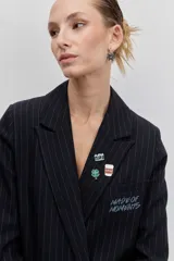 Blazer negro de rayas finas blancas, con solapas, cierre de botón frontal y broches decorativos en la solapa.