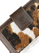 Billetera grande tipo sobre con correa de mano, confeccionada en cuero marrón con detalle central de cuero con pelo vacuno en tonos marrón, negro y blanco. Presenta cierre perimetral y solapa frontal con broche magnético y logo de búho grabado.