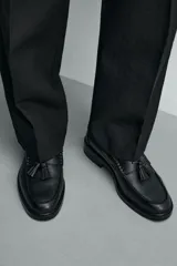 Mocasín de vestir color marrón oscuro, confeccionado en cuero, con detalle de antifaz y borlas en el empeine, horma redonda y suela plana a tono.