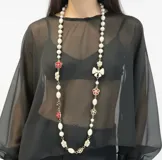 Collar largo compuesto por perlas de diferentes formas intercaladas con apliques de flores y moños de resina esmaltada en tonos rojo, negro y crema. Cuenta con cadena de alargue metálica.