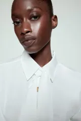Camisa blanca de cuello solapa y manga larga, con detalle de lazada en el mismo tejido en la parte frontal y cierre con botones dorados.
