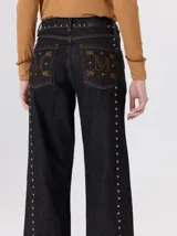 Jeans de corte ancho (wide leg) y tiro medio, confeccionados en denim de algodón negro con costuras contrastantes en color ocre. Presenta múltiples botones metálicos frontales y tachas metálicas aplicadas a lo largo de los laterales y la pretina.