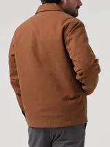 Chaqueta de canvas color marrón, con cierre frontal completo, cuello clásico y forro interior de sherpa. Presenta dos bolsillos frontales tipo parche y corte regular.