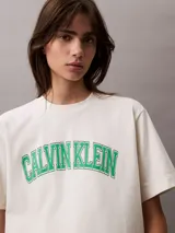 Remera de algodón color blanco hueso, corte boxy, con mangas cortas con dobladillo y logo de Calvin Klein estampado en verde. Diseño de estilo universitario.