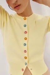 Chaqueta de punto color amarillo claro, con cuello redondo y manga corta. Presenta cierre frontal con botones de presión con diseño de flores de colores.