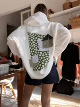 Campera tipo hoodie de corderito blanco con diseño de parches en la espalda que combinan telas estampadas con motivos florales en tonos verdes y detalles de encaje blanco.