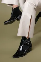 Botin de cuero negro con acabado brillante y detalle de solapa en pelo sintético, con cierre de hebilla metálica lateral y taco cuadrado.