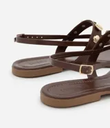Sandalia plana color beige con tiras finas y tira central decorada con colgantes metálicos dorados en forma de conchas marinas. Cuenta con cierre de hebilla ajustable en el tobillo.