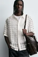 Camisa negra de corte relajado con tejido semitransparente de estructura abierta y patrón a cuadros. Presenta cuello solapa, manga corta, bolsillo plastrón en el pecho y cierre frontal de botones.