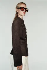 Chaqueta de gamuza marrón con cuello alto, manga larga y bajo con peplum. Cuenta con doble botonadura decorativa de botones metálicos dorados en el frente y cierre con ganchos ocultos.