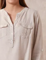 Blusa blanca de lino y rayón, con cuello a la base, escote en V y abertura con botones. Tiene bolsillos en la parte delantera y mangas largas regulables con presilla y botón.