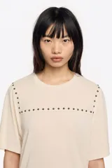 Camiseta de manga corta y escote redondo, color beige claro, con corte boxy. Presenta una hilera de tachuelas metálicas negras decorativas en la parte superior del pecho y en las costuras de los hombros.