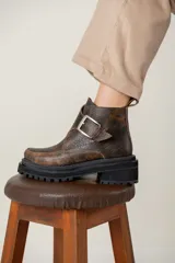 Bota corta de cuero con efecto gastado, diseño de puntera cuadrada y suela track robusta de goma negra. Presenta una hebilla metálica decorativa en el empeine y tirador en la parte trasera.