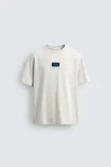 Camiseta de corte relajado, color gris jaspeado, con cuello redondo y manga corta. Presenta un parche rectangular azul marino en el centro del pecho con texto estampado en blanco y una frase adicional debajo.