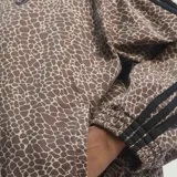 Campera deportiva Adidas modelo Firebird con cierre frontal, cuello alto y diseño estampado tipo animal print de jirafa en tonos marrones. Presenta las icónicas tres tiras de la marca en color negro a lo largo de las mangas y el logo del trébol bordado en el pecho.
