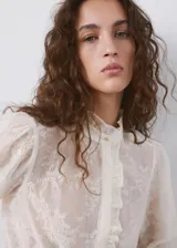 Blusa de tejido semitransparente color crudo, con diseño recto, cuello alto con volados, mangas largas abullonadas con puños abotonados y detalle de bordado floral en toda la prenda. Cuenta con cierre frontal de botones ocultos y ribete con volado en la botonera.