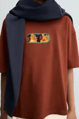 Camiseta de corte relajado color marrón, con cuello redondo y manga corta. Presenta un parche central con diseño gráfico colorido y abstracto.