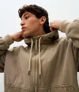 Campera trucker para hombre de sarga color beige con efecto lavado, capucha con cordón de ajuste, cierre frontal de cremallera, bolsillos tipo canguro y puños con elástico.