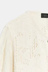 Chaqueta de cuello redondo y manga por debajo del codo con tejido combinado, color crudo, con cierre frontal con botones y detalles bordados.