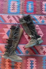 Botas de caña alta confeccionadas en cuero nobuk color chocolate, con detalle de flecos laterales en color negro, suela plana y cierre lateral.