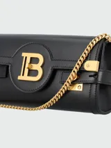 Clutch Balmain modelo B-Buzz, confeccionado en cuero negro con herrajes metálicos dorados. Presenta un diseño estructurado con cierre frontal tipo solapa, logo B metálico y detalles de correas laterales con broches grabados.