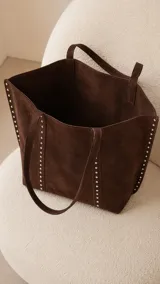 Cartera tipo tote bag de símil gamuza color marrón oscuro, de diseño amplio con detalles de tachas metálicas en los bordes y correas de hombro.