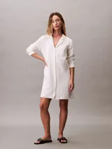 Vestido camisero corto de color blanco, confeccionado en tejido de gasa de algodón. Presenta cuello clásico, cierre frontal con botones, mangas largas y un bolsillo aplicado en el pecho.