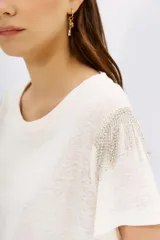 Remera blanca de lino de textura suave y liviana, con calce al cuerpo y mangas cortas. Presenta cadenas de strass decorativas en los hombros.