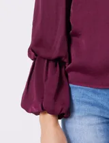 Blusa Tahari de gasa satén color violeta oscuro, con cuello alto fruncido y mangas largas abullonadas. La espalda tiene una abertura central con cierre de botón.