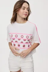 Remera blanca de manga corta, con cuello redondo, calce relajado y estampado frontal con motivos gráficos de soles y símbolos marinos en tonos rosados y bordo, además de líneas horizontales.