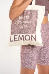 Bolsa tipo tote bag color crudo, con tiras de hombro, confeccionada en 100% algodón. Presenta un diseño gráfico frontal con estampado en tonos morados que incluye texto con las palabras "fitness, wellness, gym" y la marca "LEMON" en la parte inferior.