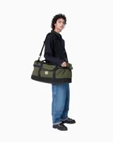 Bolso tipo duffle de la marca Carhartt, confeccionado en lona resistente de color verde militar con base y detalles en negro. Cuenta con asas de mano dobles, una correa de hombro ajustable y extraíble, un compartimento principal con cierre y un bolsillo frontal con el parche del logo de la marca.