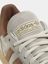 Zapatillas Adidas Handball Spezial de gamuza en tono gris claro, con las icónicas tres tiras laterales y refuerzo en el talón con textura de piel de serpiente en color marrón. Presenta suela de goma color caramelo y detalles dorados en el logo lateral y la lengüeta.