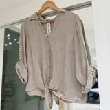 Camisa Purolino de lino color beige, con cuello camisero, escote en V con botones frontales, mangas largas con detalles calados y terminación con nudo en la parte delantera.