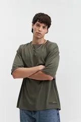 Remera oversize verde militar de manga corta y escote redondo, con estampado trasero de una cara sonriente amarilla con detalles en negro y texto "mistral 1976co".