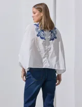Blusa blanca con bordado floral azul, escote en V, manga japonesa y cierre frontal con botones.