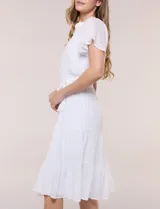 Vestido midi blanco de calce suelto y caída fluida, con escote en V, cuello a la base y apertura abotonada con detalle de puntilla. Cuenta con cintura elastizada con lazo para anudar a tono, mangas cortas con volados y falda con volantes escalonados.