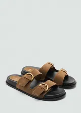 Sandalias planas de piel bovina color marrón con dos tiras en el empeine, cada una con hebilla metálica dorada ajustable. Cuenta con suela plana de color negro y punta redonda.