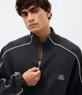 Buzo deportivo de corte boxy, color negro, con cuello alto, cierre frontal completo y mangas largas. Presenta detalles de líneas blancas a lo largo de las mangas y lettering estampado en el pecho.