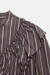 Camisa de manga larga con cuello subido, cierre frontal con botones y detalle de volantes en el pecho. Presenta un diseño a rayas verticales y puños elásticos.