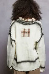 Cardigan de tejido de punto grueso color crema, con cuello tipo camisa y cierre frontal de botones florales. Presenta detalles deshilachados y teñido tipo tie-dye en tonos grises oscuros en el frente y puños. Tiene parches cuadrados de tela a cuadros en tonos tierra sobre los bolsillos delanteros.