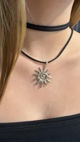 Gargantilla tipo choker compuesta por una cinta negra de gamuza y un dije metálico plateado con forma de sol con diseño en espiral.