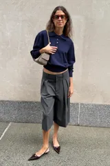 Mini bolso bandolera de piel color gris topo, con diseño fruncido, cierre mediante cordón ajustable y stopper, bolsillos exteriores laterales con solapa y botón imantado, y correa ajustable.