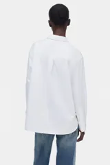 Camisa blanca oversized de manga larga, con cuello clásico, cierre frontal de botones, un bolsillo aplicado en el pecho y dobladillo redondeado.