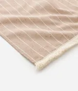 Pareo de playa rectangular de algodón en tono beige con diseño de rayas verticales blancas y acabado de flecos en los extremos.