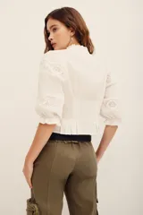 Camisa blanca de algodón con detalles de encaje broderie y transparencias, escote en V con cuello alto con volados, y mangas cortas abullonadas.