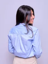 Camisa de algodón color celeste, con cuello alto, cierre frontal de botones y detalles de volados verticales a lo largo de la botonera. Presenta mangas largas con puños ajustados y terminación con volados.