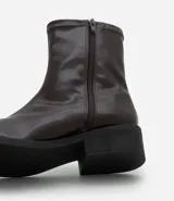 Bota tipo sock boot de caña media, confeccionada en poliuretano color marrón oscuro con acabado brillante. Presenta puntera redondeada, cierre lateral y taco bloque bajo de color negro.